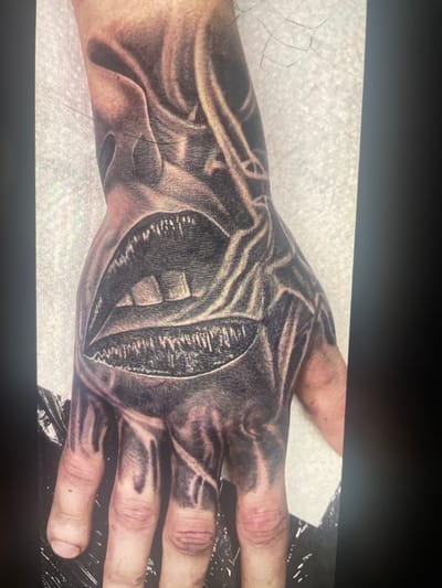 The image displays a close-up view of a black and grey tattoo on a person's hand and forearm. The tattoo depicts a pair of open lips with jagged teeth, surrounded by swirling, abstract patterns that give the impression of flames or distorted forms. The tattoo is highly detailed, with shading and line work creating a sense of depth and texture. The skin beneath the tattoo appears to be fair. The background is a textured, off-white surface, possibly a wall or fabric. There are no identifiable landmarks or elements that would suggest a specific location within the city of Paczkowo, Poland, or any other city. The image focuses solely on the artwork on the skin.
