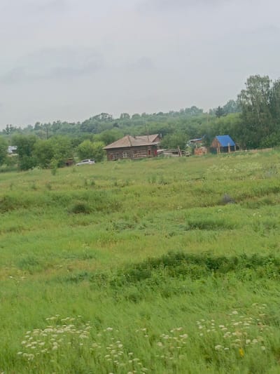 The image depicts a rural landscape in the vicinity of Акулово, Russia, characterized by rolling green hills covered in tall grass and wildflowers. A wooden log house with a red-trimmed entrance and a light-colored roof stands on the left side of a gentle slope. In front of the house, a silver SUV is parked. Further to the right, a smaller wooden structure with a bright blue roof and visible timber framing is situated. The background is dominated by lush green trees and a cloudy, overcast sky. The overall scene conveys a sense of natural tranquility and rustic living.