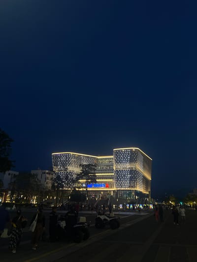 The image shows a nighttime scene in San Salvador, El Salvador, featuring a modern building adorned with lights.
**Environment:** The sky is dark blue, suggesting it is late evening or night. The area appears to be an urban setting with trees and other buildings in the background.
**Subjects and Objects:**
* A large, multi-story building with a unique architectural design is illuminated with many small white lights.
* People are visible on the street, some walking and others gathered near what appears to be a row of lit displays or kiosks.
* A police patrol with several officers and their motorcycles are on the street, possibly on patrol.
* Streetlights are scattered along the road, providing additional illumination.
**Actions and Interactions:**
* People are walking along the street and interacting with the displays.
* Police officers are present and possibly interacting with the public.
* The building stands as a central element of the scene, illuminated and attracting attention.