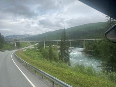 This is a photograph of a highway overpass and a fast-flowing river in a mountainous, forested region. The sky is overcast with heavy clouds. On the left, a paved highway curves to the left, bordered by a metal guardrail and green grass. In the middle ground, a long, elevated highway bridge spans across the landscape, supported by concrete pillars. Below this bridge, a wide, turbulent river flows from right to left. Another smaller pedestrian bridge crosses the river in the mid-ground. Steep, densely forested mountains rise in the background, with some patches of snow visible on the higher peaks. The image appears to be taken from a vehicle moving along the highway, as indicated by the perspective and the reflection of a car mirror in the upper right corner. The location is identified as Storforshei, Norway, suggesting this is a scenic natural landscape within or near the town. There are no people visible in the image.
