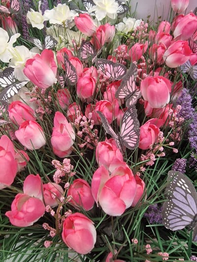 This is a close-up, eye-level, medium shot of a decorative arrangement of artificial pink and white flowers, interspersed with artificial green grass and small pink and purple floral accents. Several artificial butterflies with black and pink or black and purple patterned wings are placed among the flowers and grass. The arrangement appears to be a decorative display. The image is well-lit, with the flowers and butterflies clearly visible. There are no people present in the image. There is no information in the image to suggest a specific location in Wootton, United Kingdom.