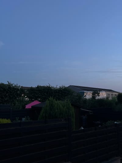 The image depicts a backyard scene at dusk in Schoonderbuken, Belgium. In the foreground, a dark wooden fence stretches across the frame. Behind the fence, a small, dark shed is visible, with a bright pink umbrella positioned in front of it. A lush green evergreen tree stands to the right of the shed. Further in the background, a dense hedge forms a natural barrier, behind which the roof and upper portion of a long building are visible. A white bus with reflective windows is parked in front of this building. The sky above is a gradient of deep blue at the top, fading to a lighter, hazy blue towards the horizon, with subtle cloud formations present. The overall lighting suggests the transition from daylight to evening.