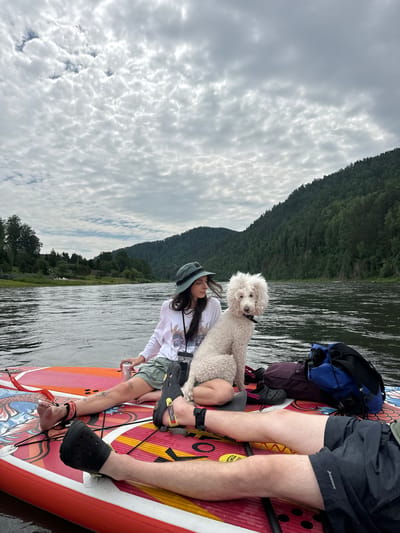 In Манский, Russia, on a river, two people and a white poodle are on a stand-up paddleboard. One person, wearing a white long-sleeved shirt and green shorts, sits with their legs crossed and holds a can. The other person, lying down, has their legs extended and is wearing black shorts. The dog sits between them, looking at the camera. The paddleboard is colorful with a dragon design. In the background, a river flows past a lush, tree-covered mountain under a cloudy sky. Two bags are placed on the paddleboard.