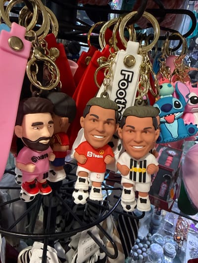 This is a close-up, eye-level shot of a display rack holding numerous keychains, likely in a souvenir shop or market stall. The keychains are predominantly in the foreground, featuring cartoonish figurines of soccer players, a Stitch character, and other colorful accessories. The soccer player keychains are dressed in miniature team jerseys and shorts, with some standing on miniature soccer balls. The figurines have exaggerated features, with large heads and smiles. Behind the keychains, the background is blurred and suggests a crowded retail environment with shelves, other merchandise, and a dark metal rack. The lighting is bright and evenly distributed, creating a slightly overexposed effect in some areas. The overall impression is one of a vibrant and busy retail display. The location is identified as Costa da Caparica, Portugal.