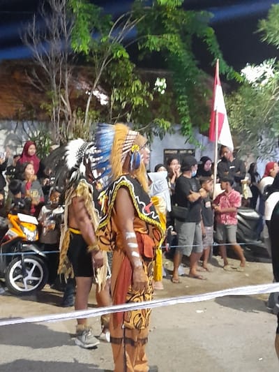 The image depicts a nighttime outdoor scene in Kedungbanteng, Indonesia, featuring two costumed individuals positioned behind a white barrier (likely a rope or tape) in the foreground, with a crowd of spectators in the background.

The central figure is a male, facing right, adorned in a large, intricate feathered headdress primarily blue, yellow, and black. He wears a golden-orange and black patterned tunic with a red sash and exhibits white paint streaks on his arms and face. To his left, partially obscured, is another male wearing a black and white feathered headdress, dark shorts, and a fur or straw-like waist garment, also with a white facial streak. Both individuals appear to be participants in a cultural or ceremonial event.

Behind the barrier, a diverse crowd of adults and children observes the scene, standing on what appears to be a paved street or public area. Many spectators are casually dressed; one woman in a red hijab is visible on the far left. In the mid-ground right, an individual holds a red and white Indonesian flag upright. A yellow and black scooter with a white front wheel is parked on the left side, behind the barrier. The background consists of dark foliage, tree branches, and a white building or wall. The scene is illuminated by artificial light, indicating nighttime.