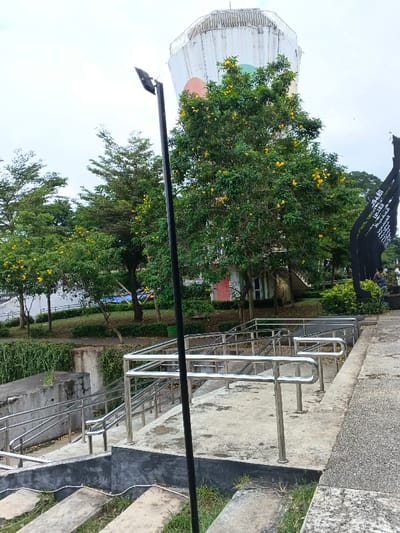 This image depicts a public park in Bien Hoa, Vietnam, featuring the Bien Hoa Water Tower. The scene is captured under an overcast, light grey sky.
In the foreground, a black lamppost with an angular light fixture stands centrally. To its left and extending downwards, rough concrete steps are partially overgrown with grass. In the midground, a multi-level concrete stairway with polished stainless steel handrails descends from the right to a lower level on the left.
The background is rich with vegetation and structures. Multiple green trees, some densely covered with vibrant yellow flowers, are distributed across the landscape. A white retaining wall, partially obscured by green vines, runs horizontally. Further back, small structures or buildings with a red door and a light green door are visible amidst the trees.
To the far right, a tall, black, angular monument or sculpture displays white text, likely Vietnamese script. Two individuals are partially visible near the base of this monument; one is seated, and the other is partially obscured behind them.
Dominating the upper background is the Bien Hoa Water Tower (Tháp Nước Biên Hòa), a tall, white, octagonal structure with a perimeter railing at its top. Its side features three large colored circular designs in orange, light green, and a darker green or blue. The ground throughout the park area is covered with grass and manicured shrubs. A green trash bin is positioned under a tree on the left side of the large, yellow-flowered tree. No explicit actions or interactions beyond static presence are discernible among the individuals.