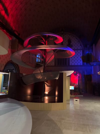 The scene unfolds within a large, high-ceilinged hall featuring vaulted architecture and intricate patterns on its ceiling and walls, indicative of a repurposed historical building, likely a former chapel or church, located in Dijon, France.
At the center of the space stands a prominent modern art installation or exhibit. This structure comprises a central vertical column from which multiple curved, ribbon-like, or helical elements extend outwards in a spiral configuration. These elements, appearing metallic or light-toned, are dramatically illuminated, displaying intense red light on their upper and left portions and blue light on their lower and right sections. The base of this central artwork integrates into a dark, cylindrical exhibit structure, which is framed by lighter-colored wooden panels.
The overall environment is bathed in striking artificial lighting. The upper left portion of the vaulted ceiling and surrounding structures are saturated with deep red light. Conversely, the far right wall, an arched niche within it, and parts of the central sculpture are illuminated with blue light. In the background to the right, an arched alcove contains a crucifix or cross sculpture, prominently highlighted by red light. A light-colored, freestanding display kiosk with a screen is visible further right, also under blue illumination. On the far left foreground, a large, translucent, white curved object, likely another exhibit component, is partially visible. The floor is composed of large, light-grey tiles laid in a regular pattern. No individuals are present within the depicted scene.