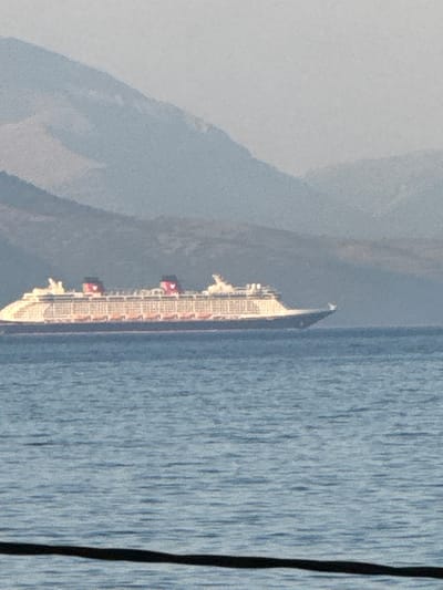 A cruise ship is sailing on a body of water. The ship is long and white with multiple decks and balconies. There are red structures visible on some of the decks. The water is calm and blue. In the background, there are hazy mountains. A dark line is visible on the right side of the image, possibly a railing or a part of the camera's viewpoint. The overall scene suggests a daytime setting, likely a clear day given the visibility of the mountains. There is no visible text in the image. The location context suggests this scene is near Meliteieis, Greece.