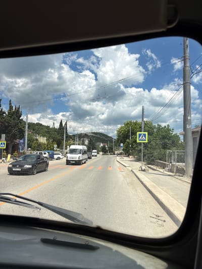 The image is taken from the interior of a vehicle, looking through the windshield. It shows a sunny day with scattered clouds. The view is of a road with several cars and vans. There is a pedestrian crossing with orange markings on the road. A yellow pedestrian crossing sign is visible on the right side of the road. Trees and buildings are visible in the background, with a hilly landscape behind them. The location is identified as Kiparisnoe, Ukraine.
