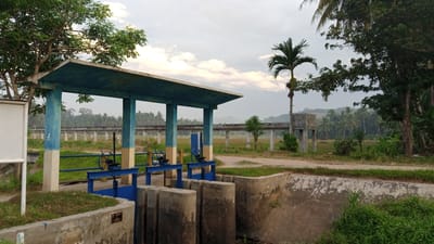 The image shows a water control structure in Blangtingkeum, Indonesia. The structure features a concrete building with a flat roof supported by pillars. Blue-painted metal gates with wheels are positioned to regulate water flow. A concrete channel runs beneath the structure. In the background, there is a long, elevated concrete walkway supported by pillars, traversing a field. Trees and vegetation are scattered around the scene. A palm tree stands prominently near the walkway. The sky is overcast.