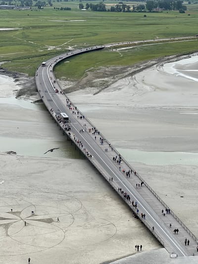 The image shows a high-angle view of a long, elevated bridge leading to Le Mont-Saint-Michel, France. The bridge is a causeway, with a road on one side and a pedestrian walkway on the other. Numerous people are walking along the bridge, some in groups and some individually. A bus is visible on the road portion of the bridge, transporting more people. The surrounding environment is a sandy tidal flat, with areas of water and green marshland visible in the distance. A bird is in flight near the bridge. On the sandy area below the bridge, there are some people drawing large geometric patterns in the sand. The sky is overcast.