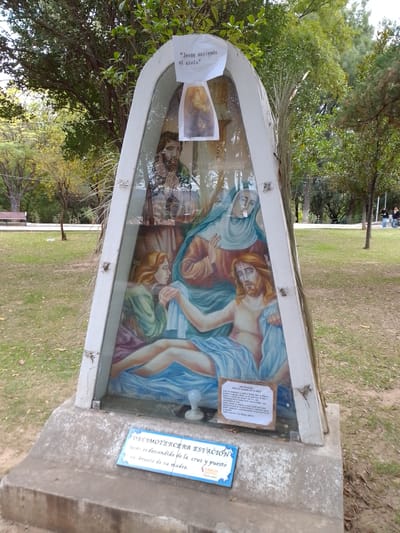 A religious monument in a park displays a painting of Jesus being taken down from the cross, cradled in the arms of his mother, Mary, and attended by others. Above this scene, a small paper bears the words "Jesus ascends to heaven" with an accompanying image. A tile at the base reads "Thirteenth Station: Jesus is taken down from the cross and placed in the arms of his mother. Tarija Capital." The monument is enclosed in a glass-fronted, white frame. The setting is a park with trees, grass, and a few people in the background. The monument is located in the city of Municipio Tarija, Bolivia.