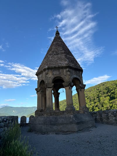 The image depicts a stone structure, possibly a bell tower or monument, with a pyramidal roof topped by a cross. The structure is octagonal in shape, supported by arches and pillars, and shows signs of age and weathering. It is situated outdoors on a clear day with a bright blue sky and scattered white clouds. The background features lush green hills, a body of water, and distant mountains. A low stone wall surrounds the area. The foreground is covered in gravel. The image does not show any people or actions. The structure is located in Shalikiantkari, Georgia, as identified by the context provided.