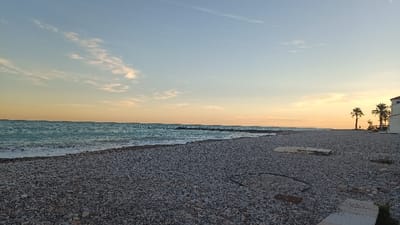 The image presents a serene coastal landscape at El Grau de Moncofa, Spain, captured during what appears to be either sunrise or sunset. The sky is a prominent feature, transitioning from a light blue at the top to a warm gradient of soft orange and yellow hues near the horizon, with delicate, wispy white clouds scattered across it.
The foreground is dominated by a broad expanse of a dark pebble or shingle beach, which fills the lower half of the frame. Scattered on the pebbles are several light-colored, rectangular flat objects and a dark, curved line that resembles a pipe or cable.
On the left side, the calm, turquoise-blue sea meets the shore with small, gentle waves breaking into white foam. A low, dark breakwater or a line of rocky structures is visible in the middle distance, extending horizontally across the water, separating the immediate shoreline from the wider ocean.
To the right, two distinct palm trees stand tall, their dark silhouettes contrasting against the lighter sky. Adjacent to the palm trees, a white building with an orange or terracotta-tiled roof is partially visible, suggesting a coastal dwelling or structure. A wooden fence or railing is also discernible near the base of the palm trees, leading towards the building.
The scene is devoid of people or direct activity, conveying a peaceful and tranquil atmosphere characteristic of the golden hour. No visible text is present in the image.