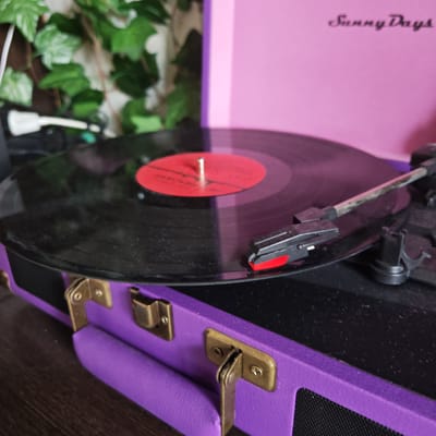 The image shows a purple and pink "Sunny Days" record player with a black vinyl record on the turntable. The record has a red label. The stylus is engaged with the record. The record player has brass-colored latches and a black mesh speaker. In the background, there are green vines. The location is not discernable and the city of Ketoshi, Russia cannot be confirmed from the image. There are no people visible in the image.