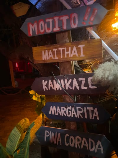 Five signs for cocktails are arranged in a row. The signs are made of wood and painted with the names of the cocktails in white lettering: "Mojito," "Maithai," "Kamikaze," "Margarita," and "Pina Corada." The signs are situated among greenery and appear to be part of a bar or restaurant setting. The location is Baan Don Sai, Thailand.