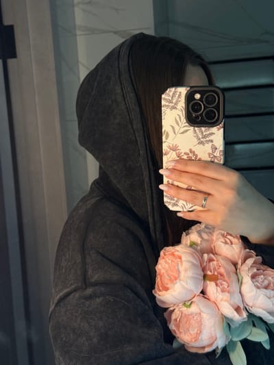 A person is depicted taking a mirror selfie indoors in Tliustenkhabl', Russia. The individual, primarily obscured, wears a dark grey, possibly distressed, hooded sweatshirt. Their face is largely hidden by a smartphone held in their right hand, though long, dark hair is visible. The smartphone, likely an iPhone model with a prominent triple-lens camera system, is encased in a light-colored cover featuring a botanical pattern of leaves, flowers, and butterflies over vertical stripes. The person's right hand exhibits manicured fingernails with light polish and white tips, and a silver-toned ring is worn on the ring finger. A bouquet of several light pink artificial peonies with green leaves is held in the crook of the left arm, resting against the chest. The background, visible in the mirror reflection, shows vertical elements suggesting wall paneling or tiles with a light and dark grey or white and black color scheme.