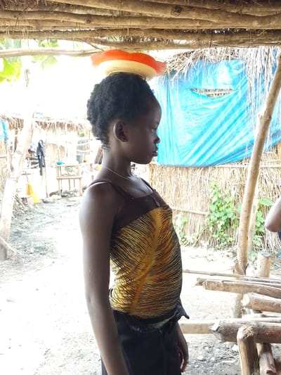 A young African girl stands in profile, facing right, with a circular, orange and white object balanced on her head. She has dark, curly hair and is wearing a brown and yellow patterned tank top and dark pants. Her expression is neutral as she looks off into the distance.
The setting appears to be outdoors, under a makeshift shelter constructed with wooden poles and a thatched roof. In the background, there is a blue tarp covering a wall made of dried vegetation. Further back, more rustic structures and some miscellaneous items, including a table and buckets, are visible. The ground is rough and dusty.
The scene suggests a rural or informal settlement in Otulu, Nigeria. The lighting indicates it could be daytime, possibly with bright sunlight filtering through the shelter. There is no text visible in the image. The overall impression is of a candid moment, possibly depicting a daily task or activity.