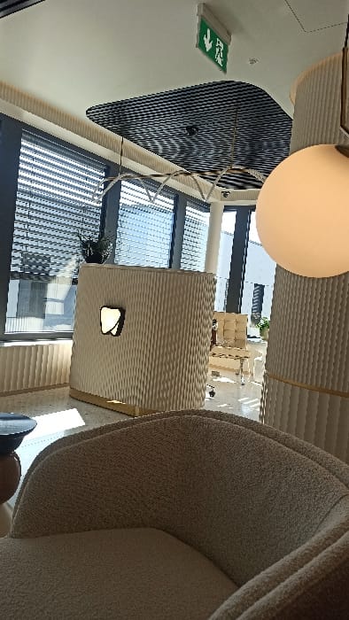 This image depicts a modern, unoccupied reception or waiting area located in Bertrange, Luxembourg. In the foreground, the back and seat of a light-brown, textured upholstered armchair are partially visible on the bottom right. Centrally positioned is a light-toned reception desk featuring a vertically ribbed surface and a gold-colored base. The desk displays an illuminated, tooth-shaped emblem on its front and a small dark green potted plant on its top surface. Behind the desk, large windows equipped with horizontal blinds overlook an exterior building. To the right, a prominent spherical white pendant lamp hangs, partially obscuring a light-colored, vertically ribbed cylindrical column. Beyond the reception desk, through an architectural opening, a cream-colored armchair and a small table are visible in an adjacent waiting space. The ceiling is white, incorporating a dark, textured, possibly acoustic, panel. A green emergency exit sign with a downward arrow and a running figure is mounted on the white ceiling. Linear, geometric pendant light fixtures are also suspended from the ceiling. The floor is light-colored and speckled. No individuals are present, and no actions or interactions are depicted.