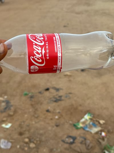 An empty, clear plastic Coca-Cola bottle lies on sandy, dirt ground littered with debris and a small green plant. The bottle is positioned horizontally and is partially held by a person's fingers, which are visible on the left side of the frame. The Coca-Cola branding is prominent on the red label, indicating "Original Taste" and "Carbonated Soft Drink." Text on the label also mentions "Great Taste - Less Sugar" and "50 cl." In the background, the ground is uneven and speckled with small stones. Further in the distance, more scattered trash is visible. The image appears to be taken outdoors in a dry, potentially urban or semi-urban environment.