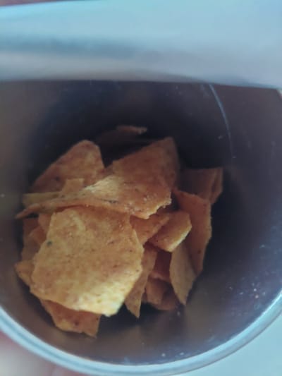 An overhead shot shows a close-up view of tortilla chips piled inside a metal container. The chips are golden yellow and appear seasoned with small dark flecks. The container is dark gray on the inside and has a metallic rim. The lighting suggests an indoor environment. There is no identifiable location in Jelutung, Indonesia depicted in this image.