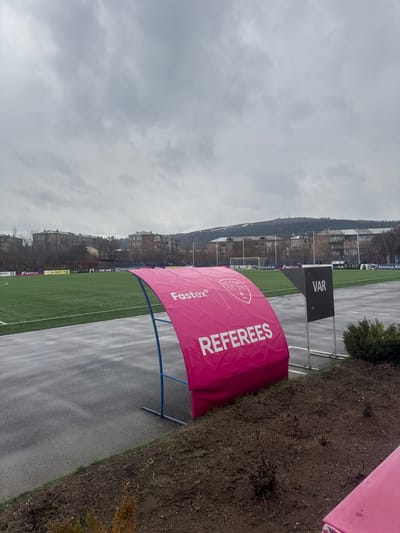 The image shows a view of a soccer field in Charentsavan, Armenia, on a cloudy day. A pink "REFEREES" tent is visible near the edge of the field, along with a sign that says "VAR." The background includes buildings and hills, typical of the area. The field appears to be artificial turf, and a wet pathway runs alongside it. Sponsorship banners line the field, with visible logos including "FASTBANK" and "YO HEALTH." The overall impression is of a match day setup, possibly during inclement weather.