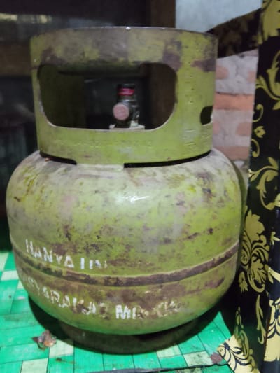 A close-up, eye-level, medium shot captures a weathered, green LPG gas cylinder positioned indoors. The cylinder, showing signs of rust and wear, rests on a green mat with a diamond pattern. A portion of a dark cloth with a yellow floral pattern is visible on the right side of the frame, with a brick wall in the background. The cylinder has white lettering that reads "HANYA UNTUK" and "SYARAKAT MISKIN" which translates to "ONLY FOR" and "POOR COMMUNITY" in Indonesian. The location is the city of Merbopuntong, Indonesia.