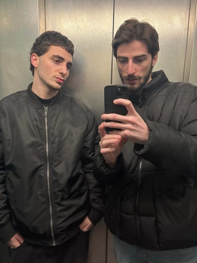 Two men are captured in a mirrored image within an elevator. One man, wearing a black bomber jacket, is positioned at the bottom. Above, the other man wears a puffy black jacket and holds a black smartphone, seemingly taking the picture. The mirrored reflection creates the illusion of two separate individuals. The elevator walls are visible in the background, providing context to the setting. Both men gaze towards the camera, engaging the viewer.