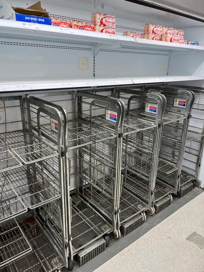 The image shows a stack of metal shopping carts inside a building at the coordinates -27.556, 152.923 in Middle Park, Australia. The carts are made of wire mesh and are stacked on top of each other. The carts are empty and are sitting in a aisle of a grocery store. The area is well-lit and clean.  The image captures the carts from a low-angle perspective, showcasing the stacked structure. The carts are arranged in an orderly manner, suggesting that they are ready for use. The scene is devoid of people or other objects, except for a few boxes of refrigerated beverages stacked in a refrigerator on the left side of the image. The background shows a white tiled floor with a black drain.  The image is taken from an angle, allowing the viewer to see the full height of the stacked shopping carts.