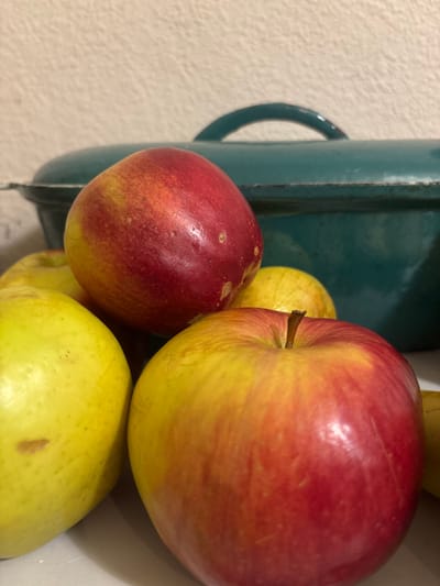 A close-up, low-angle shot shows a pile of apples in front of a teal Dutch oven. The apples are mostly red and yellow, with some green ones visible. The Dutch oven is dark teal and appears to be made of cast iron. The background is a textured, off-white wall. The scene is lit from above and to the left, casting soft shadows. There are no people or animals visible in the image. This image does not provide any information about the location in the city of Svkh Maslovskiy 1 Otdeleniye, Russia.