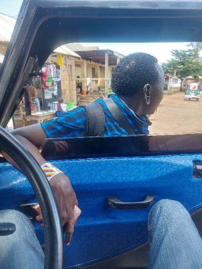 The image depicts a street scene in Kimamba, Tanzania, viewed from inside a vehicle. The interior of the vehicle is predominantly blue with a textured finish, featuring a black steering wheel on the left and a black door handle. The driver's dark-skinned left arm, adorned with a multi-colored beaded bracelet (white, yellow, orange, red, blue, green), rests on the steering wheel while holding a small, light brown rectangular object. The driver's denim-clad legs are partially visible in the lower foreground.
Seated to the driver's right, a second individual, with dark skin and short, dark hair, is seen from behind, looking out the window to the right. This person wears a blue patterned short-sleeved shirt and a dark strap, possibly from a backpack, crosses their shoulder. An earring is visible on their right ear.
The exterior scene reveals an unpaved street or open area. On the left, a rudimentary shop or stall is visible, displaying various items, including clothing hanging from a structure. A person wearing a light shirt and blue jeans is seated at a table near this stall. Further back, another individual in a light-colored top is present near green containers. Buildings of simple construction, some with corrugated metal roofs, line the street. One building features a small porch with wooden supports. In the distant background to the right, a light-colored, small three-wheeled vehicle, possibly a tuk-tuk, is parked or in motion. Sparse trees and vegetation are visible under a clear or hazy sky.