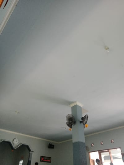 The image depicts the interior of a large room in Kawungluwuk, Indonesia, captured from a low-angle perspective looking upwards. The ceiling is light blue with visible patches of lighter grey, suggesting varied illumination or paint tones. A white, bare lightbulb fixture is visible on the ceiling in the upper right quadrant. A prominent light blue and grey rectangular pillar stands in the middle-right foreground, extending from the floor to the ceiling, topped with a white capital. Three black wall-mounted fans with visible yellow mounting brackets are affixed to this pillar: two on the left side and one on the right.
The walls are predominantly grey, featuring white trim along the ceiling line. On the left wall, a decorative archway or niche, also grey with white trim, frames a circular white wall clock displaying approximately 10:10. Below this archway, a rectangular electronic display board with red digital readouts is mounted on the wall. Above the archway, a dark rectangular object, possibly a speaker, is affixed high on the wall.
In the background on the right, a large rectangular opening, possibly a window or doorway with a visible wooden frame, reveals an outdoor setting or another room. Three small, dark, rectangular wall decorations are mounted above this opening. The lower edge of the image partially shows the heads of two individuals near the framed opening on the right, one appearing to wear a light-colored shirt. No specific actions or interactions are discernible from their partial presence. The lighting appears to be natural, casting soft shadows, particularly from the ceiling trim.