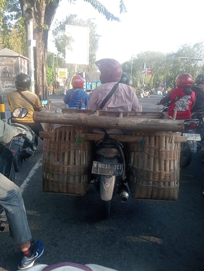 In the city of Banjarkemantren, Indonesia, a photograph captures a street scene with multiple motorcycles. The primary focus is on a motorcycle with a large, elaborate bamboo basket structure attached to its rear, extending outwards. This basket appears to be carrying goods or supplies. Behind this motorcycle, another rider is visible, wearing a red helmet and a red hooded jacket with a graphic design. To the left, a person in a yellow t-shirt and black pants, wearing a black helmet, is on a motorcycle. Further in the background, a traffic light and trees line the street, with more motorcycles and a blue truck visible in the distance. The ground is paved asphalt.