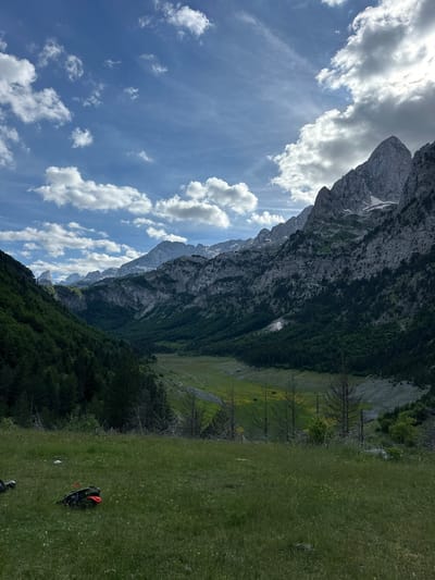 The image depicts a panoramic view of a mountainous valley in Shalë, Albania. In the foreground, a grassy meadow is dotted with small yellow wildflowers. Two backpacks, one black with an orange element and another black, are placed on the grass in the lower left portion of the frame. The middle ground reveals a lush green valley floor, possibly a meadow, with scattered dead trees. This valley is flanked by steep, densely forested mountainsides on either side. The background is dominated by a dramatic mountain range with jagged, rocky peaks, some of which are partially covered in snow. The sky above is a vibrant blue, interspersed with scattered, fluffy white clouds, and sunlight breaks through in some areas, casting rays on the mountains. The overall scene conveys a sense of wild, natural beauty and rugged terrain.