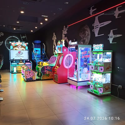 The image depicts an indoor arcade or entertainment center. The scene is dominated by various arcade machines, many of which are illuminated with neon and LED lights in vibrant colors such as blue, pink, green, and yellow. Prominently featured are several claw machine games, a racing simulator game with a pirate-themed marquee, and other video game cabinets. Decorative elements on the walls include silhouette cutouts of Star Wars-themed spacecraft and figures, as well as a large circular graphic resembling a planet. The floor is tiled in a light beige color, with colorful light reflections from the machines. A watermark in the bottom right corner indicates the date "24.07.2026" and time "10:16". The location is identified as Modugno, Italy. There are no people visible in the image.