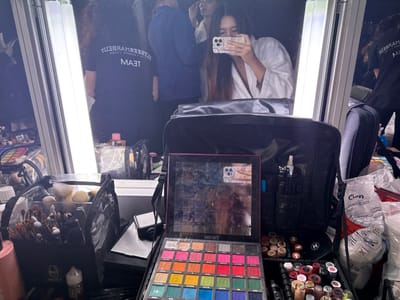 The image displays a professional makeup station located in a backstage dressing room environment in Issy-les-Moulineaux, France. The central focus is a large mirror flanked vertically by two bright fluorescent light tubes.

In the immediate foreground, an open makeup palette, predominantly black, features a grid of numerous vibrant, multi-colored eyeshadows, some exhibiting clear signs of prior use. To the left of the palette, a black bag or container is filled with an extensive collection of makeup brushes of various sizes and types. Adjacent to this, on the right, a black, open professional makeup artist's case or trolley bag is visible, organized into compartments holding an assortment of lipsticks, bottles, and other cosmetic tools. A plastic bag, partially labeled "Cien," is also present on the far right.

The mirror reflects a woman with long, dark hair, wearing a white bathrobe, holding a silver-colored smartphone with a triple-lens camera array, actively taking a photograph or selfie. Reflected to her right, another individual is seen from behind, wearing a black t-shirt with inverted white text reading "TEAM" and "MARBERAM BEAUTY." The reflections also reveal additional makeup products and organizational items on a counter behind the primary setup, indicating a busy and functional preparation area.