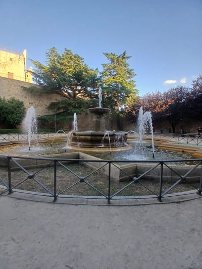A fountain in a courtyard in Ávila, Spain, is the central focus of the image. The fountain has multiple tiers and streams of water spraying into a basin. Behind the fountain, a stone wall and trees are visible, with a building peeking out from behind the trees. A black metal fence encloses the fountain area. The sky is clear blue. In the background, two people can be seen walking.