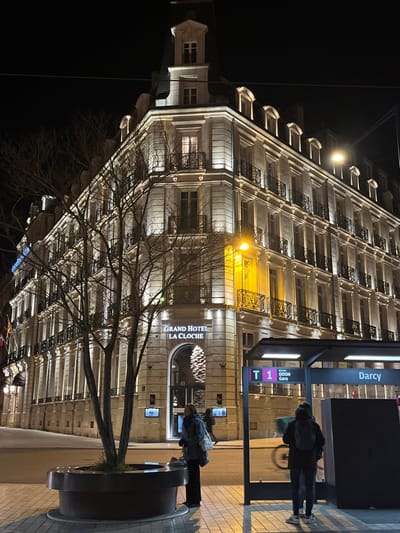 A tall, well-lit building, the Grand Hotel La Cloche, dominates the nighttime view. Bare tree branches reach up in front of it. A bus stop is visible, with "Darcy" and "T 1" markings. Pedestrians wait at the stop and near a circular planter. Streetlights illuminate the scene. The image is oriented sideways.