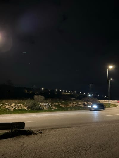 A landscape-oriented nighttime photograph captures a road scene, likely in Anatoliki Attiki, Greece. The image is taken from a slightly elevated position alongside a two-lane road, which appears to have a somewhat wet or reflective asphalt surface, possibly from recent rain. A dark-colored car is visible on the right side of the road, moving away from the viewer, its bright headlights creating a luminous glow on the asphalt ahead.

Along the left side of the road, a long line of streetlights extends into the distance, casting distinct circles of light onto the ground and illuminating the surrounding terrain. This area consists of a rocky embankment covered with sparse green vegetation and shrubs, suggesting a semi-rural or natural setting. A dark metal guardrail is visible along the immediate edge of the road in the foreground. The sky, which occupies the majority of the left side of the image, is completely dark, indicating a clear night. Some minor light artifacts or lens flare are visible in the darker areas of the sky. The scene is devoid of people or any discernible text. The strong contrast between the bright artificial lights and the deep darkness of the night sky and unlit areas is a prominent feature.