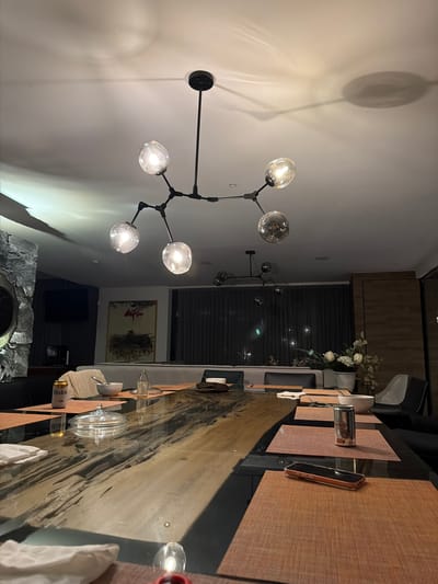 The image depicts an interior dining space. A long wooden table, set with place mats, plates, glasses, and napkins, dominates the foreground. A modern chandelier with multiple globe lights hangs above the table. In the background, there are window blinds, a cabinet, a piece of art, a television, and a stone feature. The scene is lit with artificial light. The location is Santa Ana Yancuitlalpan (Chilhuacán), Mexico. There are no people visible.