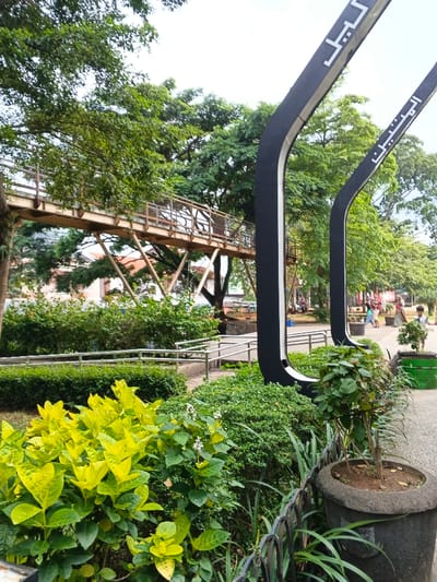The image captures a vibrant public park or green space, filled with lush vegetation and architectural elements. In the mid-ground and background, an elevated pedestrian walkway or bridge, constructed from metal with railings, stretches across the scene, supported by a lattice of metal beams. Numerous large trees with dense green foliage dominate the upper and background areas, suggesting a mature and well-established park environment.
Two prominent, tall, dark-colored, curved structures with white Arabic script are a central feature on the right side of the image. The left-most of these structures displays the word "الليل" (Al-Layl), meaning "The Night". The right-most structure shows the word "المنيب" (Al-Muneeb), meaning "The Penitent" or "The Repentant". These structures appear to be decorative arches, gateway elements, or part of a larger artistic installation.
Below these large structures, a paved path is visible, bordered by low metal railings, suggesting a ramp or accessible pathway. Further back, several people are scattered across a more open, paved area, casually walking or standing, indicating a public gathering spot within the park. Some appear to be adults, possibly with children.
In the immediate foreground, a variety of plants and bushes are densely arranged. On the left, a large bush with bright, almost chartreuse-yellow leaves stands out. To its right, a darker green bush with small white flowers adds texture. A black metal fence segment is visible along the very bottom edge. In the bottom right corner, a dark, heavy concrete planter pot holds a small plant with slender, grass-like leaves and some broader, green leaves.
The lighting suggests it is daytime, possibly late morning or afternoon, under a bright but somewhat overcast or diffused sky, as there are no harsh shadows. The overall atmosphere is tranquil and inviting, typical of a well-maintained urban park.