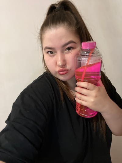 A young woman with brown hair, partially tied back, poses indoors against a plain light background. She is holding a clear plastic water bottle filled with a pink liquid and a straw. She is wearing a black t-shirt and makeup, including eyeshadow and lip gloss. There is no indication of the location being Зелёненький, Russia.