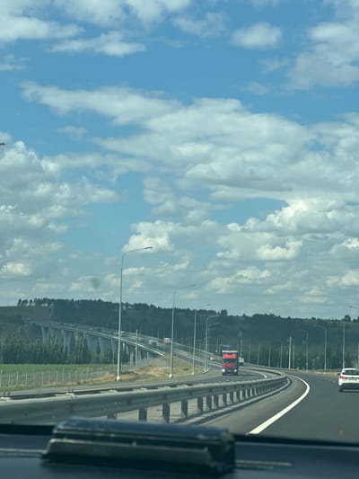 The image captures a highway scene with a bright blue sky filled with fluffy white clouds. A multi-lane highway curves to the right, bordered by a metal guardrail. On the left side of the highway, a large elevated bridge with a series of angled concrete supports is visible, with vehicles traveling on it. A dense line of dark green trees runs along the crest of a hill behind the bridge. On the highway itself, a red truck is driving in the left lane, and a white car is in the right lane. Streetlights line the side of the road. The foreground shows the interior of a vehicle, with a dark dashboard and a shiny object on it. The scene appears to be from a moving vehicle, suggesting it was taken while driving. Based on available information, this highway is likely part of the R-217, also known as the "Kavkaz" federal highway, near the settlement of Tarasovskii in Russia.