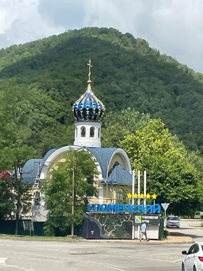 An Eastern Orthodox church with a blue and gold dome stands in a lush, green, mountainous area. A sign with blue lettering spelling out "ТЮМЕНСКИЙ" (Tyumenskiy) and "ТУАПСИНСКИЙ РАЙОН" (Tuapsinsky District) is visible in front of the church, with yellow stars above it. The sign also features a map. A person in a light blue shirt and beige pants walks away from the sign. A white car is parked on the right side of the road, and another white car is partially visible in the foreground. The sky is cloudy.