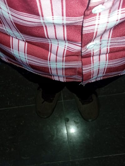 The image shows a close-up view of a person's torso and legs, from the waist down, wearing a red and white plaid shirt, dark trousers, and light brown sneakers. The shirt has two visible white buttons. The scene is indoors, with a dark tiled floor visible beneath the person's feet. The lighting is dim, suggesting it could be evening or an indoor setting with low illumination. There are no visible signs of weather, specific time of day, or emotional expression. The setting appears to be a simple, everyday environment, possibly a home or public space. The image focuses on the clothing and footwear of the subject. There is no visible text in the image.