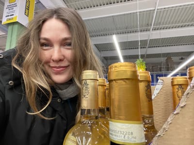 A close-up, upward-angled shot captures a woman and a display of bottled beverages within what appears to be a large retail store. The image is oriented with the top of the woman's head towards the bottom of the frame, and bottles hanging from what looks like a cardboard display box above her.
The central subject is a woman with light brown or blonde hair, parted in the middle and framing her face. She has light-colored eyes and her lips are closed in a slight, neutral smile. She is wearing a black puffer jacket over a grey hooded top. Her face is positioned in the lower right quadrant of the image.
Above and slightly to the left of her, several bottles containing a light golden liquid are prominent. They are either suspended or part of an overhead display, with their bases facing downwards towards the viewer. The bottles have golden foil-wrapped necks and dark caps. One bottle clearly shows part of a label with the text "000232208" and "WXST 0.75L", indicating a volume of 0.75 liters. Another partial label shows "KJ /". The bottles appear to be packaged within a torn cardboard box, visible in the upper left.
The background reveals the interior of a commercial establishment, likely a supermarket or warehouse store. The ceiling features white, ribbed panels and bright fluorescent strip lighting. A green, spherical object, possibly a hanging decoration or artificial plant, is visible dangling from the ceiling structure. In the lower right corner, a white sign with the prominent "METRO" logo is visible. Below the logo, some Cyrillic text is present, with legible parts reading "МОБИЛЬНОЕ ПРИЛОЖЕНИЕ" (Mobile Application). The overall lighting suggests an indoor setting during operating hours.