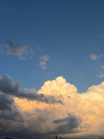 The image depicts a dramatic sky filled with clouds in the city of Slobozia, Moldova. The upper portion of the frame is dominated by a clear, deep blue sky, gradually transitioning to a more intense blue towards the top. Scattered white and grey clouds drift across this expanse. The lower half of the image is a spectacle of large, billowing cumulus clouds. These clouds are illuminated from within by warm, golden sunlight, creating a striking contrast with the darker, shadowed undersides of some clouds on the left side of the frame. The overall impression is of a dynamic and visually striking celestial display. The image does not contain any people, subjects, or specific objects that would indicate actions or interactions. The environment is solely the sky, with faint hints of silhouettes of trees and possibly a rooftop structure at the very bottom edge of the frame, suggesting a ground-level perspective from within Slobozia.