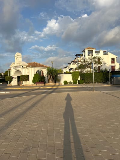 The image depicts a street scene in Vera Playa, Spain, during what appears to be late afternoon or early evening, evidenced by the long shadows cast by the setting sun. In the foreground, a paved walkway of interlocking grey bricks stretches across the frame, with a prominent shadow of a person elongated across it.
To the left of the image is a small, white, single-story building with a red tiled roof, featuring an arched entrance. Above the arch, text reads "LOS JARDINES DE NUEVA VERA." A tall, slender lamppost stands to the right of this building. In front of the building, manicured green bushes and a palm tree are visible, along with a red bench.
Further back and to the right, a multi-story white building with multiple balconies and red-shuttered windows rises. This building exhibits a modern architectural style. A hedge separates the property from the street. In the middle ground, a street sign and a tall pole are present. On the far right, a grey garbage bin is visible. The sky above is a mix of blue with scattered white and grey clouds, indicating a dynamic weather pattern. The overall impression is one of a well-maintained residential or complex area.