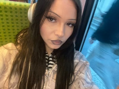 A front-facing selfie captures a young individual, likely female, with long, dark hair accented by lighter streaks, wearing light-colored over-ear headphones. The subject's face features prominent winged eyeliner, dark lipstick, a septum piercing, and multiple studs piercing the left eyebrow. They are dressed in a black and white striped top beneath a light-toned, possibly patterned, zippered hooded jacket, featuring a stylized cross-shaped zipper pull.

The subject is seated inside a vehicle, indicated by a green, geometrically patterned fabric seat visible on the left side of the frame. To the right, a window reveals a blurred exterior environment. Through the window, a partially visible figure in dark upper clothing and light lower garments is captured in motion, suggesting movement of either the vehicle or the passerby. Below this figure, a segment of a platform edge with distinct tactile paving (raised circular bumps) is visible, consistent with a train station or bus stop.

This scene is located within a public transport vehicle, likely a train or bus, at or in the vicinity of a station in Enghien-les-Bains, France.