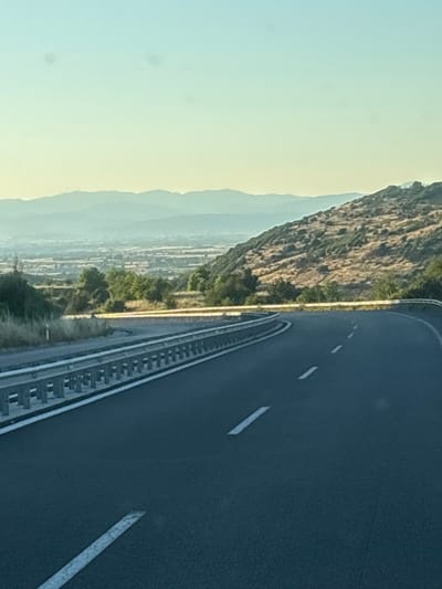 The image shows a curved asphalt road with white dashed and solid lane markings. On the left, a metal guardrail runs along the side of the road, separating it from a dry, grassy embankment. Beyond the embankment, trees and a distant valley with buildings and more hills are visible under a clear, pale blue sky. The road appears to be in a rural or semi-rural area, possibly a highway on the outskirts of the city of Avra, Greece. The lighting suggests it is either early morning or late afternoon. There are no people or vehicles in the image.